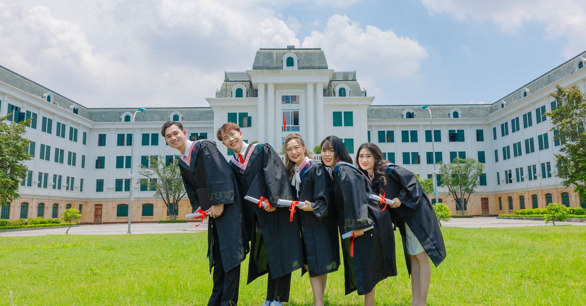 Group of Asian graduates celebrating with diplomas in front of a university building on a sunny day.